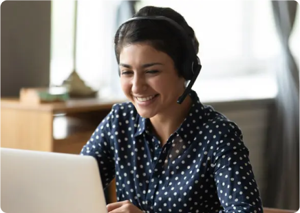 A woman happily using a laptop with a headset on, indicating she is engaged in a conversation or online communication.